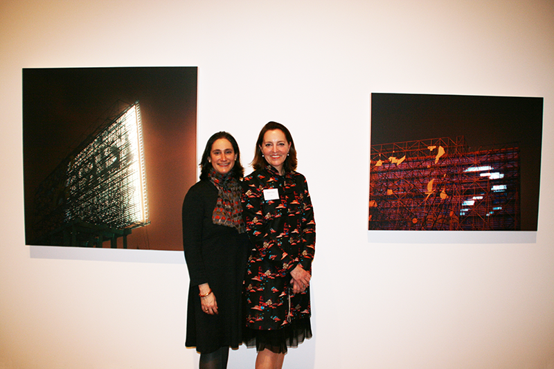 A color photograph of two light-skinned women. They each wear a knee-length dress and have shoulder length brown hair; they stand close together to smile for the camera. Behind them on a white gallery wall are two large photographic prints of the backs of stadium lights.